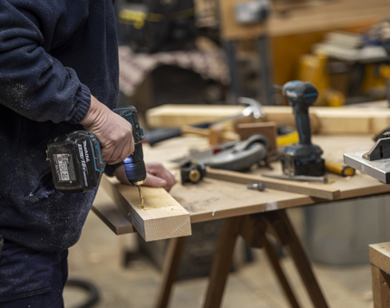 A maintenance team member standing at a work bench holding a drill to a piece of wood. 