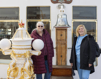 Two women stand next to The Royal Yacht Britannia's Bell and decorative binnacle on the Verandah Deck. 