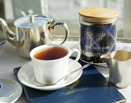 A cup of tea is on a table with a silver teapot and milk jug and a container of Pekoe Tea Royal Yacht Blend tea.