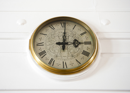 A gold‑rimmed wall clock with Roman numerals hangs on a white wall, its ornate black hands showing the time at three o’clock.