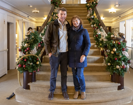 A man and a woman stand in front of Britannia's Grand Staircase which is decorated with festive garlands on the banisters. 