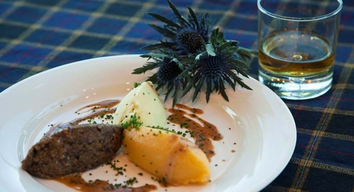 A plate of haggis, neeps and tatties and a glass of whisky on a tartan tablecloth. 