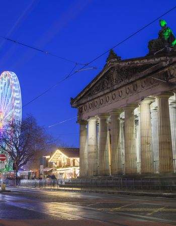 The National Galleries of Scotland on the Mound in Edinburgh at Christmas. There is an illuminated big wheel and German Market in the image.