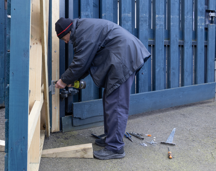 A man from the Facilities team attaching a hinge to a blue wooden gate in the compound on the Quayside. 