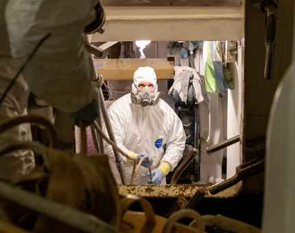 Two men from Maintenance wearing protective suits and masks are spray painting in side a room. 