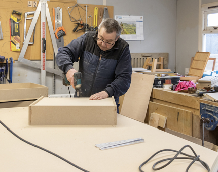 A man uses a drill to put together a wooden drawer for a podium. 