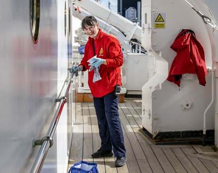A smiling woman from housekeeping wearing a red jacket polishing a handrail outside on deck at Britannia. 