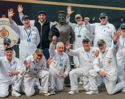 Two rows of Yotties are in a group waving at the camera, while standing next to a bronze statue of a former Yottie. 