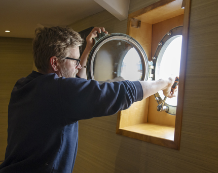A man is using a wrench to tighten fittings on a porthole in a cabin