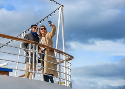 Two people stand on a ship’s deck taking a selfie, dressed in coats and sunglasses. White railings, string lights, and a bright sky with scattered clouds create a relaxed, scenic atmosphere.