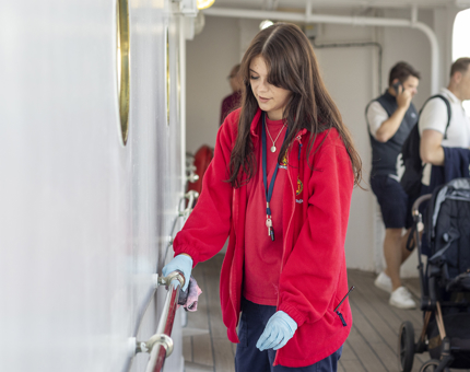 Lady with dark brown hair and wearing a bright red t-shirt and fleece polishing the silver handrails along the deck.