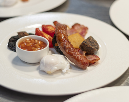 A plate with a Scottish breakfast containing sausage, egg, baked beans, tomato, mushroom, haggis and bacon. 
