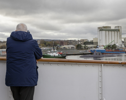 A visitor looking out over the water to a submarine being towed into the port. 