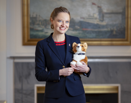 A woman standing in the Drawing Room holding a corgi plush toy. 