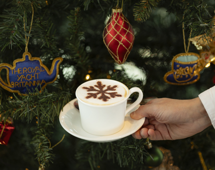 A hand holding a cappuccino with a chocolate snowflake stencil on top of it. There is a Christmas tree in the background. 