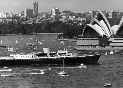 Britannia sails through Sydney Harbour in a black‑and‑white scene, surrounded by small boats. The Sydney Opera House rises on the right, with the city skyline behind it, giving the moment a historic, ceremonial feel.