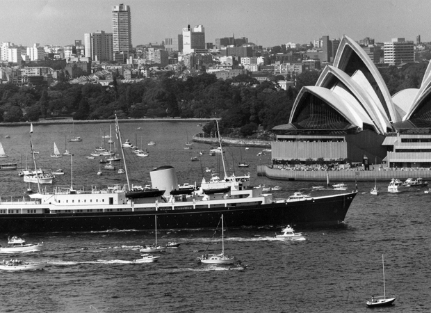 Britannia sails through Sydney Harbour in a black‑and‑white scene, surrounded by small boats. The Sydney Opera House rises on the right, with the city skyline behind it, giving the moment a historic, ceremonial feel.
