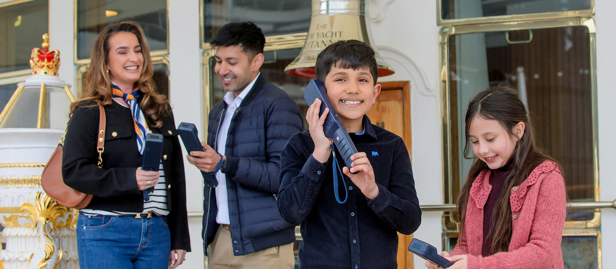 A family listening to an audio guide handset next to Britannia's Bell and binnacle.