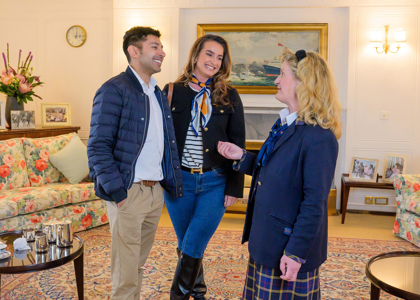 Three people stand chatting in an elegant lounge furnished with floral sofas, a patterned rug, and a coffee table set with a silver tea service. Framed photographs, a ship painting, wall lamps, and a clock create a formal, historic atmosphere as one person gestures mid‑conversation.