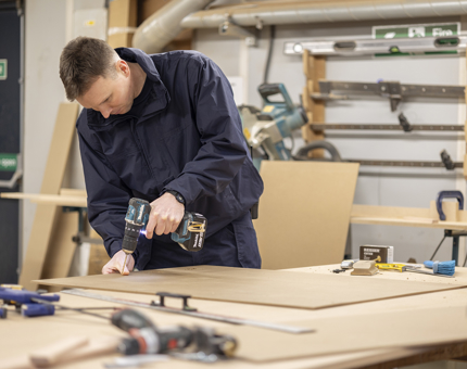 A Maintenance team member in the Joiners' Workshop making storage. He is standing at the bench, holding a drill, drilling a piece of wood. 