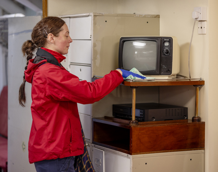 A Housekeeper wearing a red fleece is wiping down a stand that has a tv on top of it. 