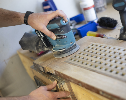 A close up of a hand holding an electric sander, sanding a scupper board. 