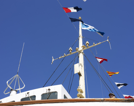 A view looking up to the Bridge with dress flags flying above the ship, the sky is blue. 