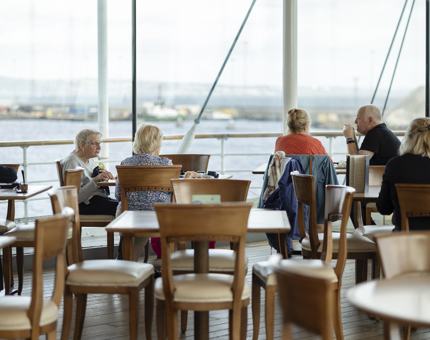 Visitors sitting at the tables in the Royal Deck Tearoom. There is a view of the Port of Leith through the large picture windows. 