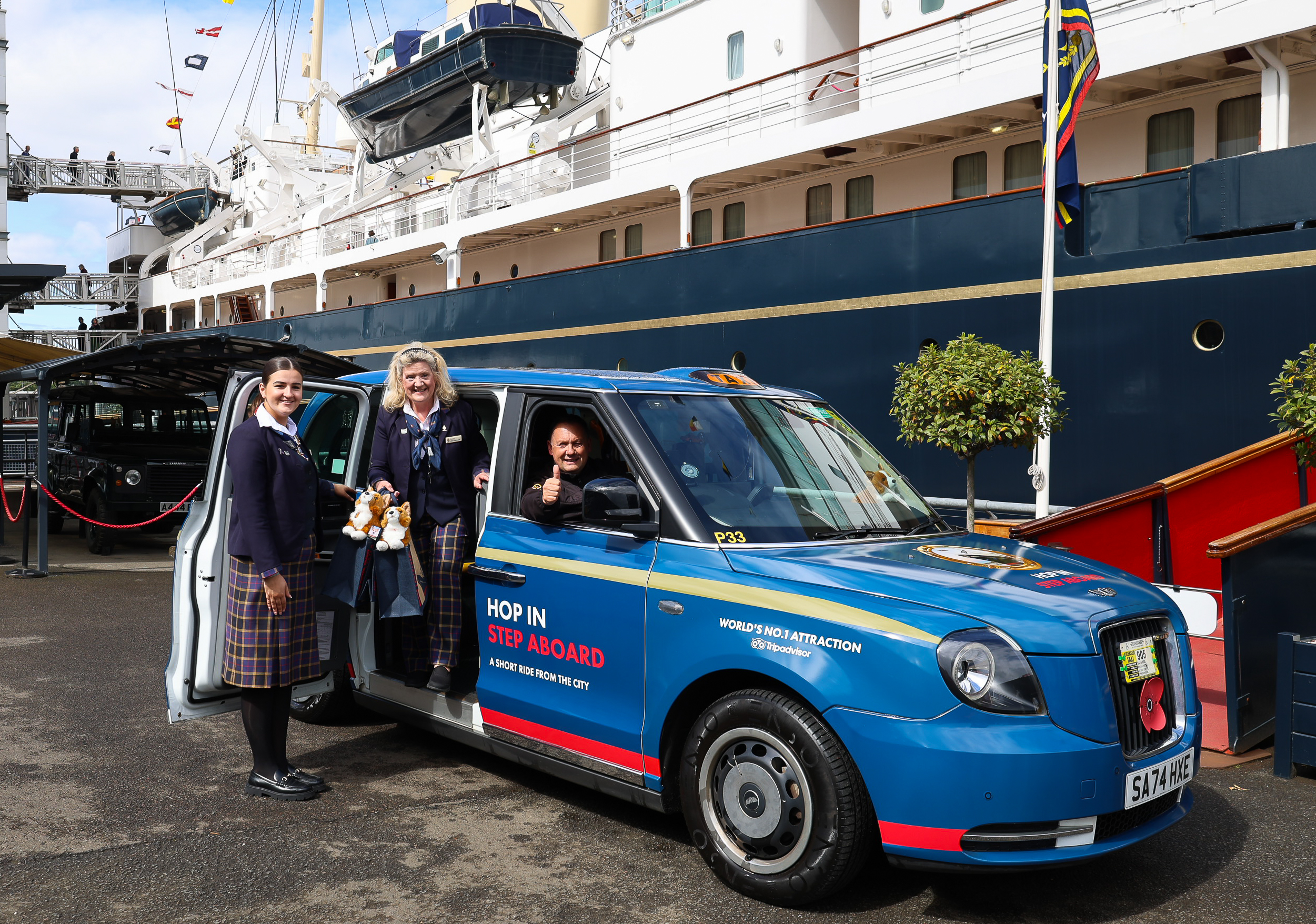 Two visitors pose beside a blue taxi parked in front of a large ship. One visitor sits inside the taxi giving a thumbs‑up, while two others stand outside wearing plaid skirts and navy blazers. The taxi displays the text “Hop In Step Aboard” and promotes a VisitScotland attraction.