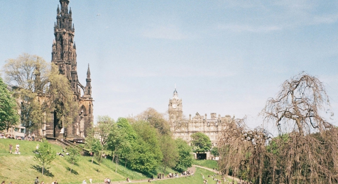 A picture of Princes Street Gardens in Edinburgh. There are lots of people sitting on the grass enjoying the sunshine. The Scott Monument is on the left hand side of the picture. 