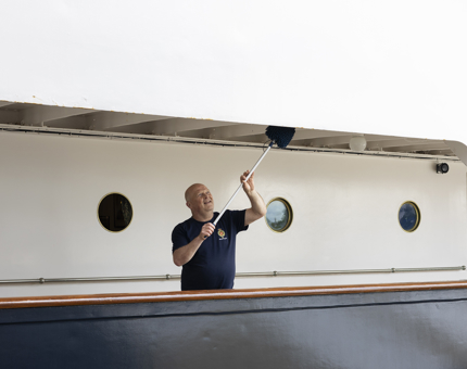 A Facilities Officer is holding a duster, cleaning deck heads on the port side of Britannia. 