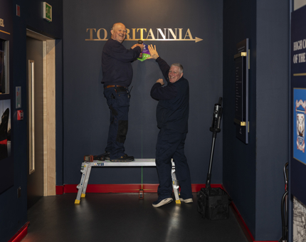 Two Facilities Officers are attaching a small sign to a blue wall in Britannia's Visitor Centre.