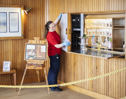 In the Sun Lounge, a Housekeeper is wiping down a wood panelled drinks cabinet. 