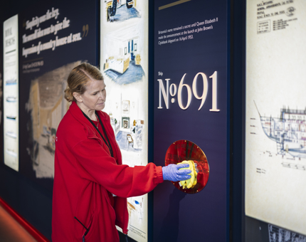 A Housekeeper is using a yellow cloth, polishing a brass plaque in Britannia's Visitor Centre. 