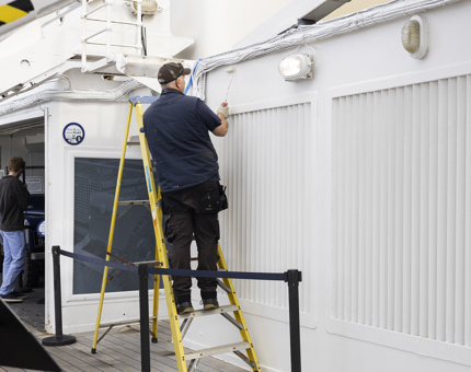 A maintenance man is standing on a ladder using a paint roller to apply a coat of paint. 