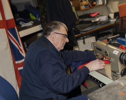 A man is sitting at a sewing machine sewing dress flags for Britannia. 