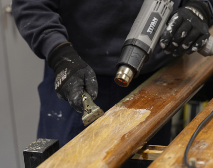 A close up of hands holding a heat gun and scraper, removing varnish from a wooden handrail. 
