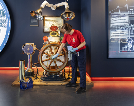 A Housekeeper is in the Visitor Centre and is polishing the brass on a ship's wheel. 