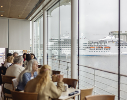 Visitors sitting at tables next to the windows in the Royal Deck Tearoom. There is a view of a cruise ship docked in the Port of Leith.