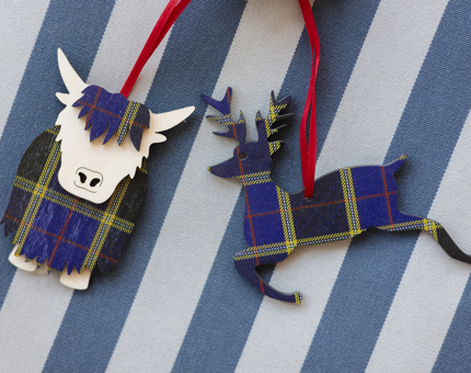 Two wooden hanging decorations. They are a blue tartan pattern, one is a deer, the other is a highland cow. 