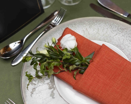 A close up of a table setting in Fingal's Ballroom. There is a green table cloth with white plates and an orange napkin folded with flowers coming out of it.