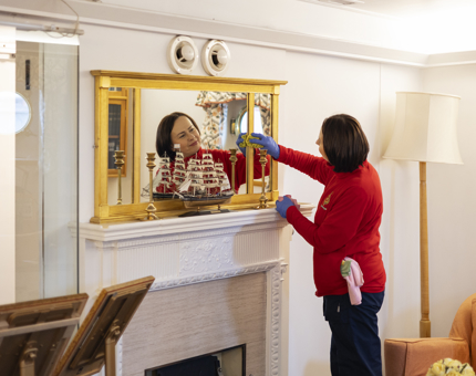 A Housekeeper is cleaning a mirror above a fireplace aboard Britannia. You can see her reflection in the mirror. 