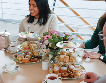 Guests are dining in the Royal Deck Tearoom aboard The Royal Yacht Britannia. There are cake stands and cups of tea on the table. 