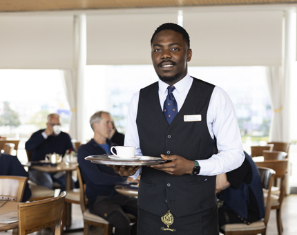 A Hospitality Server is holding a tray with a coffee on it in the Royal Deck Tearoom. 
