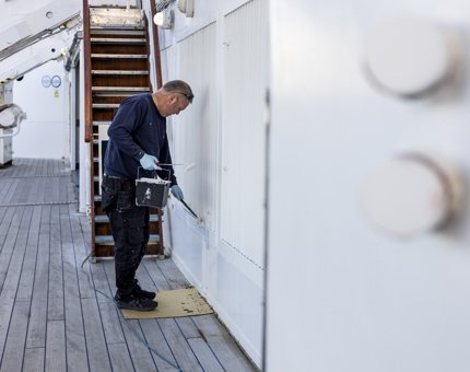 A maintenance man is standing on Britannia's deck, he is holding a paint roller and bucket of paint, applying it to the wall. 