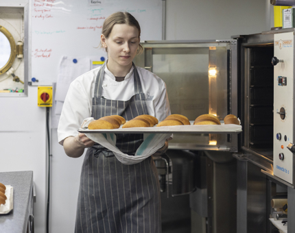 A chef is in the Galley, she is holding a tray of buns, fresh out of the oven. 