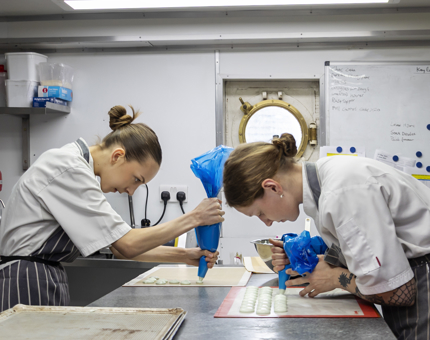 Two Chefs in the Galley holding piping bags, piping macaron batter onto a tray.