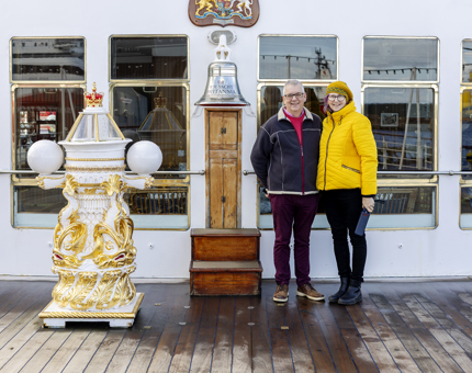 Two visitors posing for a photo on Britannia's Verandah Deck next to the Bell and decorative binnacle. 