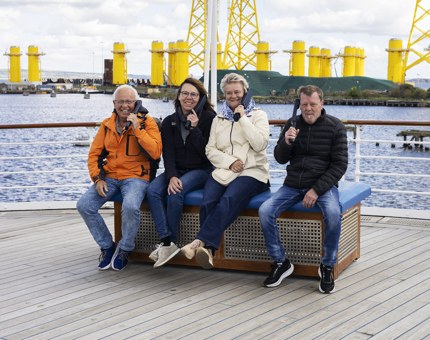 Four people sitting on a bench aboard Britannia int he Port of Leith. There are large yellow structure in the background. 