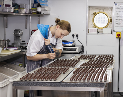 A Chef is in the Galley, she is piping caramel into small chocolates to be made into petit fours. 